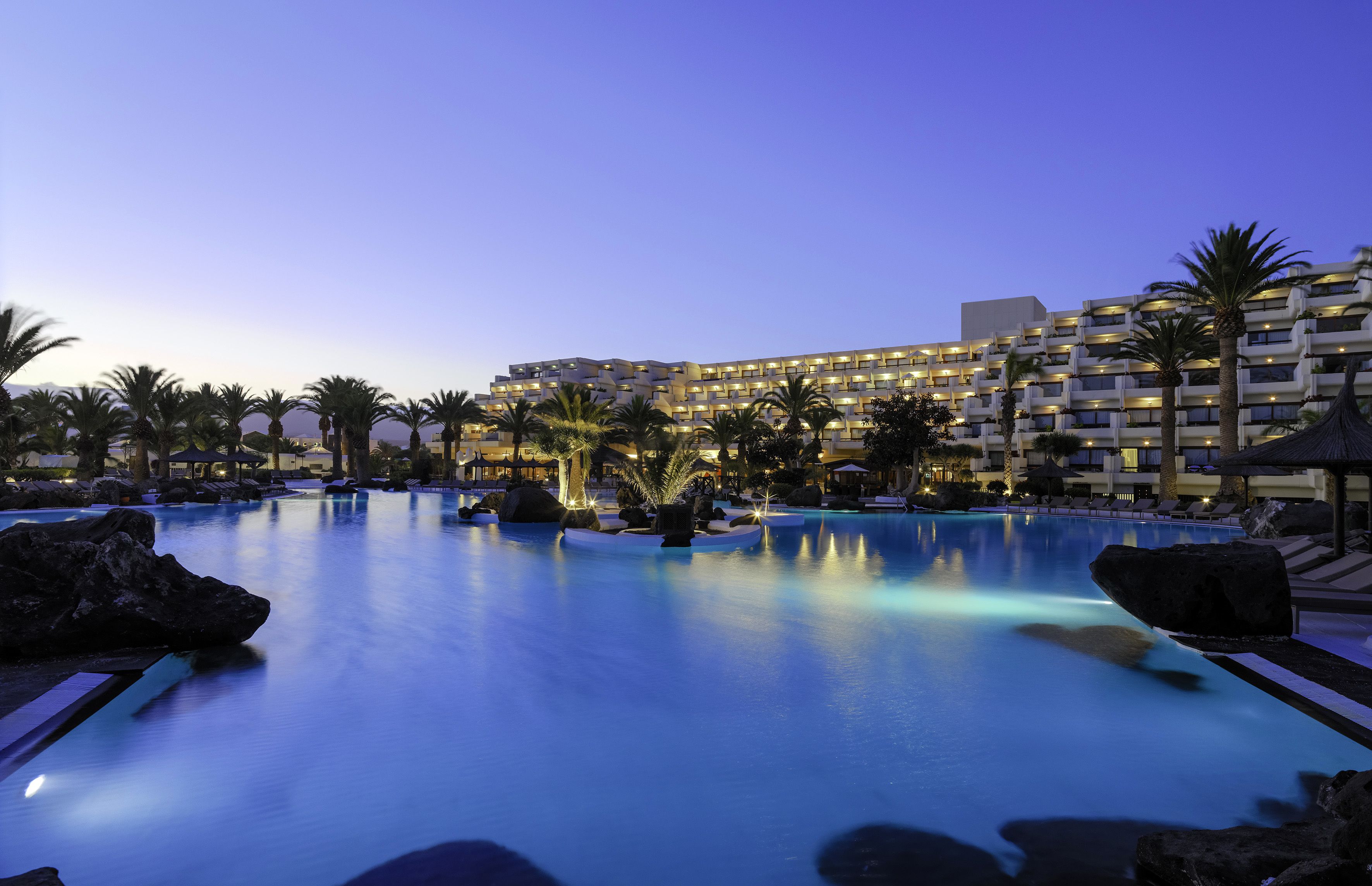 Un hotel con una gran piscina, palmeras y un edificio moderno con varios balcones.