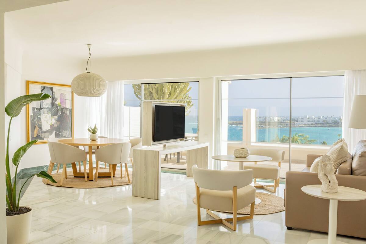 A modern living room with a TV, dining table, and seating area, featuring large windows with a city view.