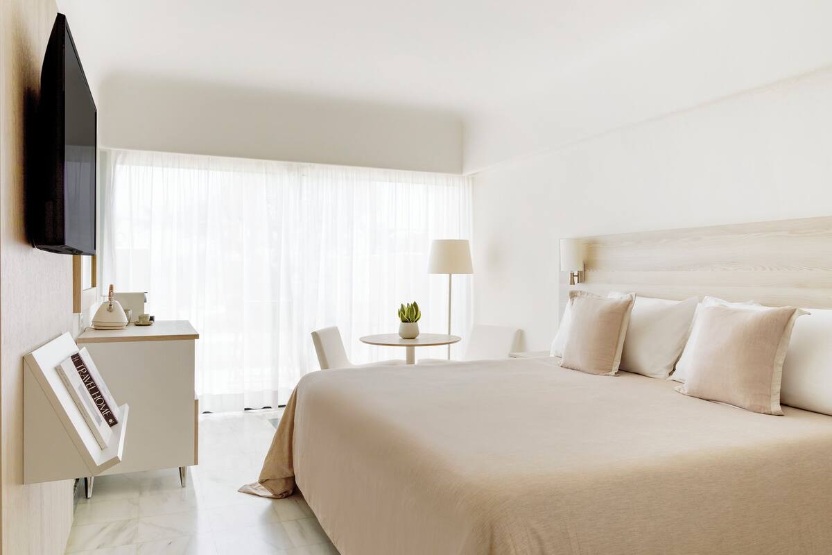 A hotel room with a large bed, a desk with a lamp, a television, and a window with sheer curtains.