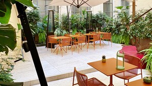 Un patio interior con mesas y sillas, rodeado de exuberante vegetación y grandes sombrillas que ofrecen sombra.