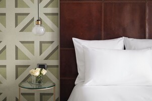 A neatly made bed with white linens, a glass side table with a vase of white flowers, and a geometric patterned wall.
