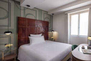 A hotel room with a large bed, a small table with a cup, a chair, a lamp, and a window with curtains.