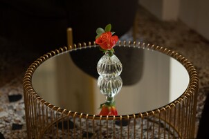 Una mesa redonda de cristal con base de alambre dorado, un florero transparente con una sola rosa roja y el reflejo de la rosa en la superficie de la mesa.