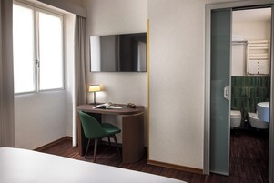 A hotel room with a bed, a desk with a chair, a TV mounted on the wall, and a bathroom visible through an open door.
