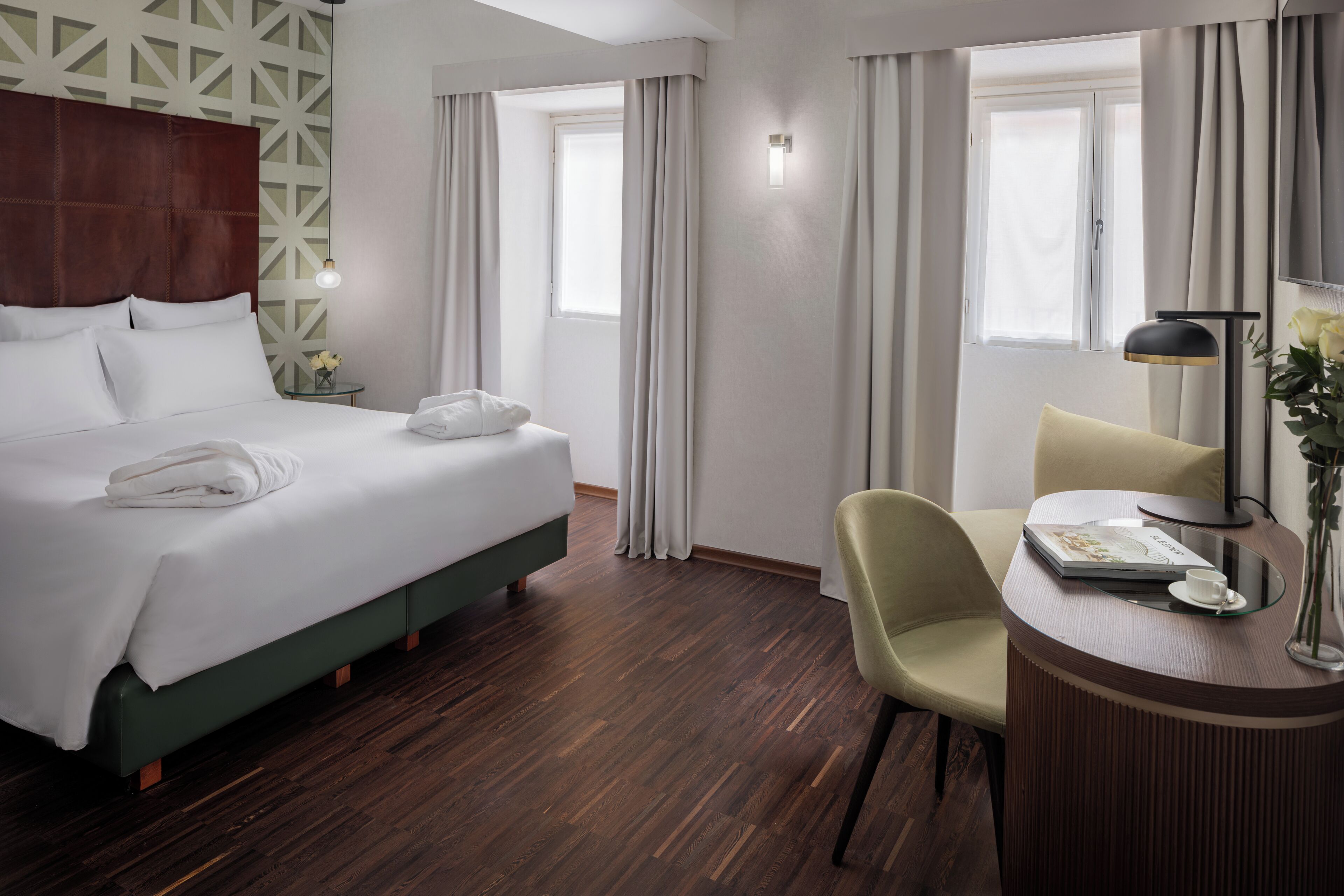 A hotel room with a large bed, a desk with a chair, a small table with a cup and a book, and a vase with flowers.