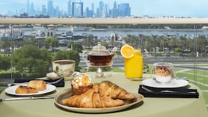 Een ontbijttafel met croissants, muffins, een kop thee, een glas sinaasappelsap en een yoghurt parfait, met uitzicht op een stadsgezicht en een jachthaven.