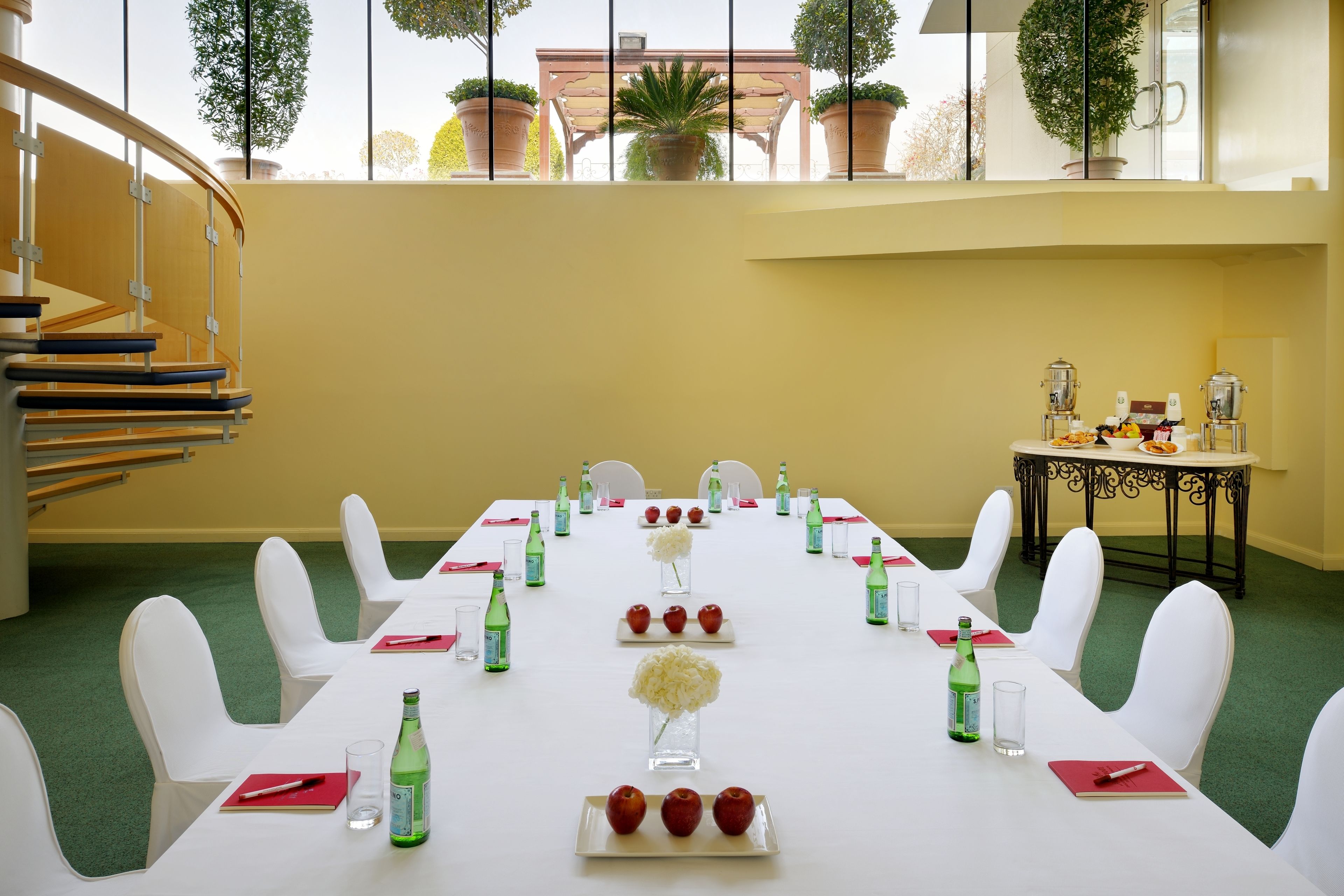 Sala de conferências com uma longa mesa preparada para uma reunião, cadeiras brancas e uma escadaria ao fundo.