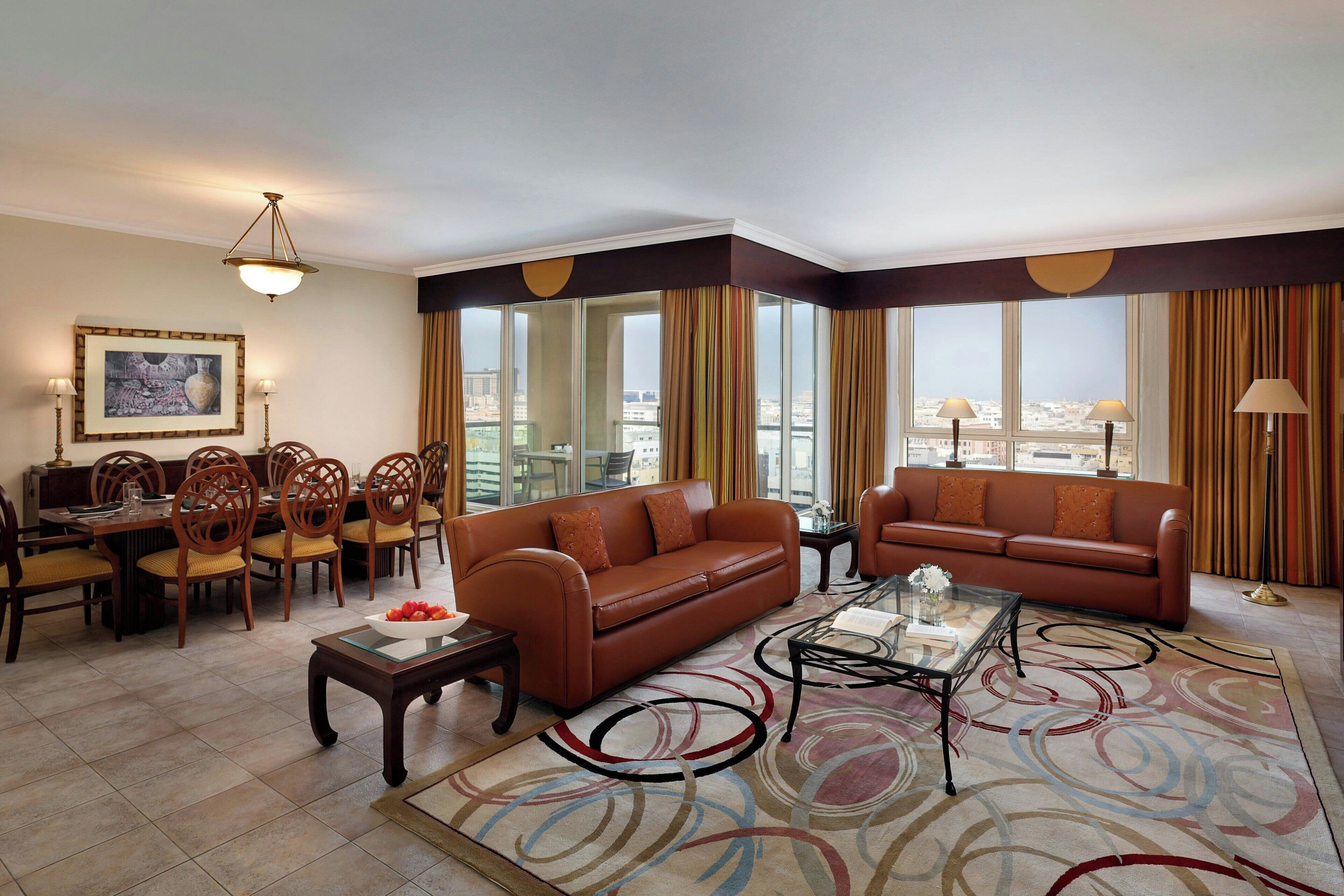 Habitación de hotel con sofá de cuero marrón, una mesa de centro de vidrio, zona de comedor con sillas de madera y ventanas grandes con cortinas.