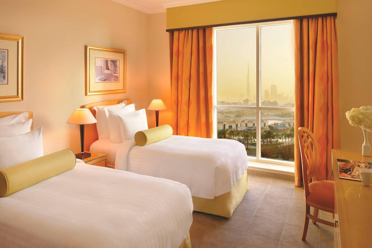Une chambre d’hôtel avec deux lits, un bureau, une chaise, une fenêtre donnant sur la ville et des œuvres d’art encadrées accrochées aux murs.