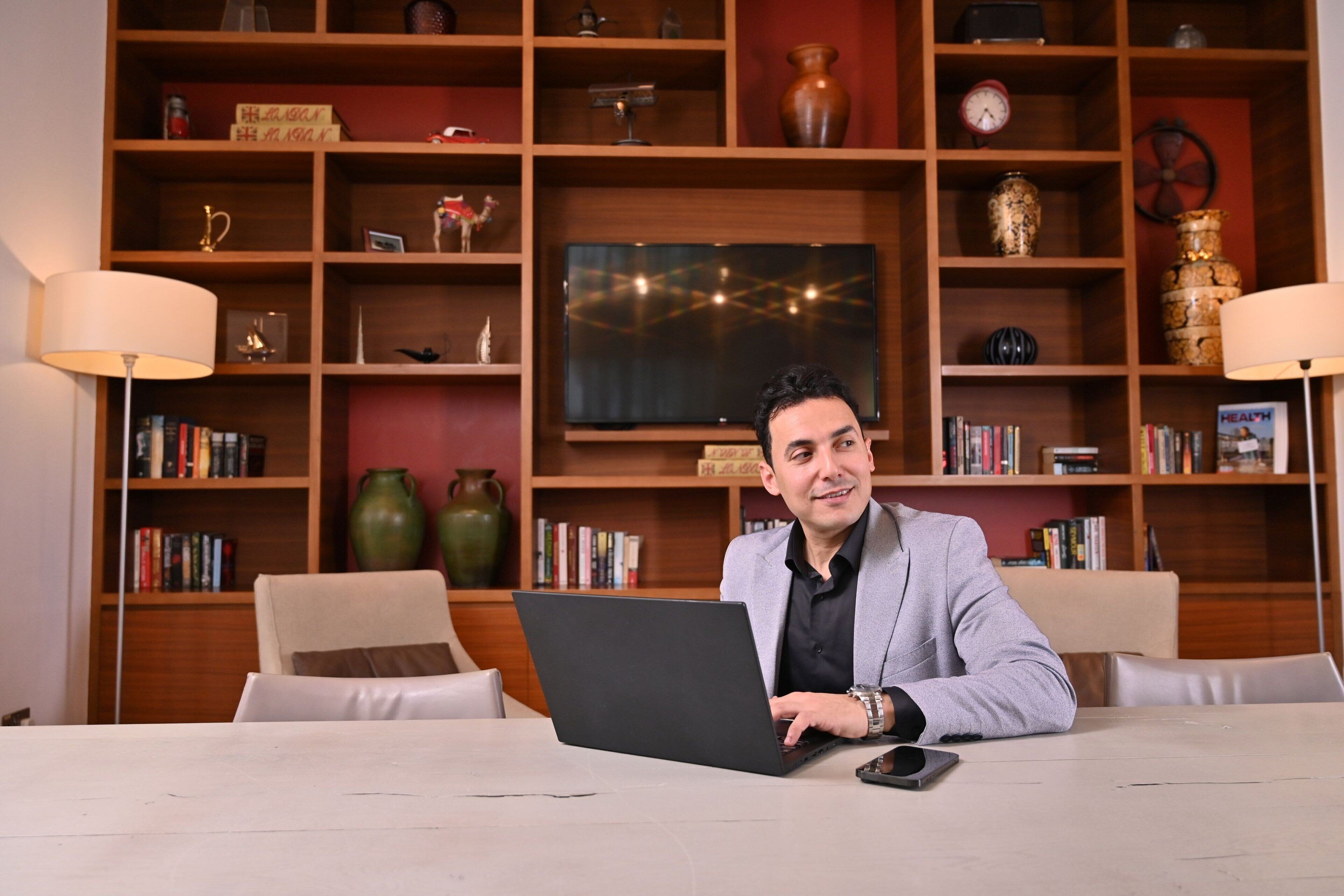 A man sitting at a table with a laptop in a room with wooden shelves containing books, decorative items, and a television.