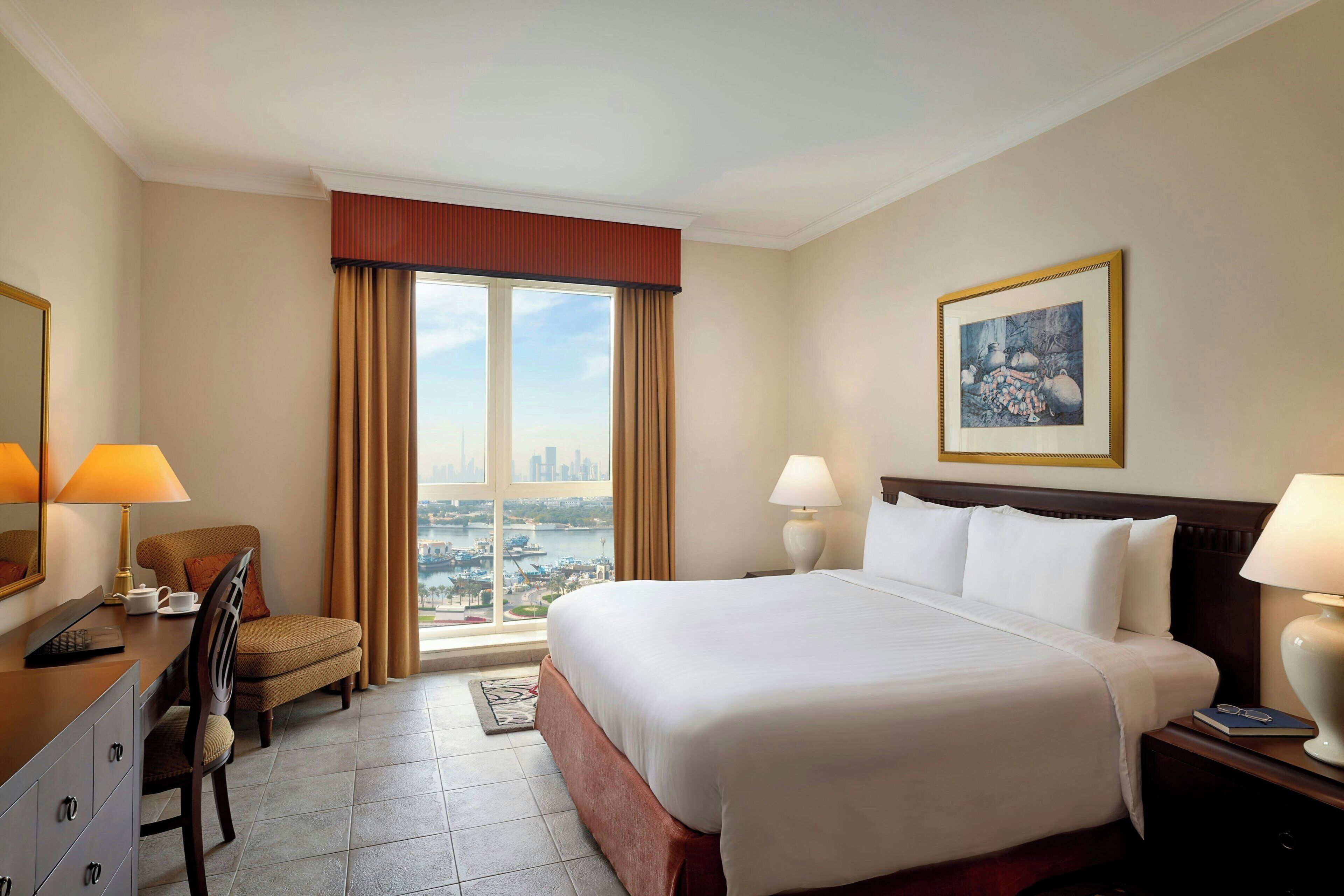 A hotel room with a large bed, two bedside lamps, a chair, a desk with a lamp, a framed picture on the wall, and a view of a cityscape through a large window.
