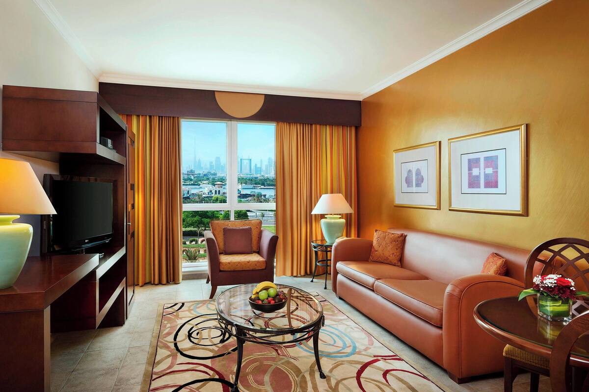 Une chambre d’hôtel moderne dotée d’une grande fenêtre donnant sur la ville, d’un canapé confortable, d’une télévision et d’un coin repas avec une table et des chaises.