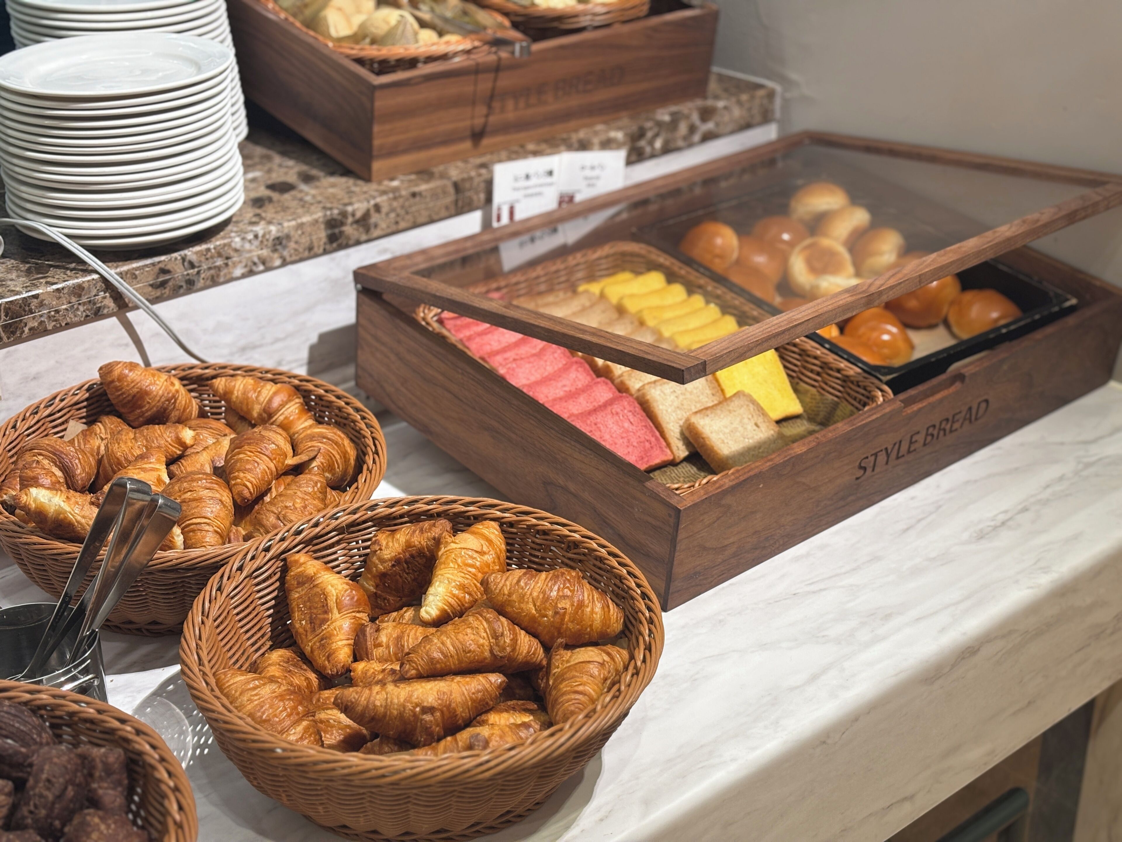A buffet with various bread and pastries, including croissants and baguettes, displayed in wicker baskets and wooden boxes.