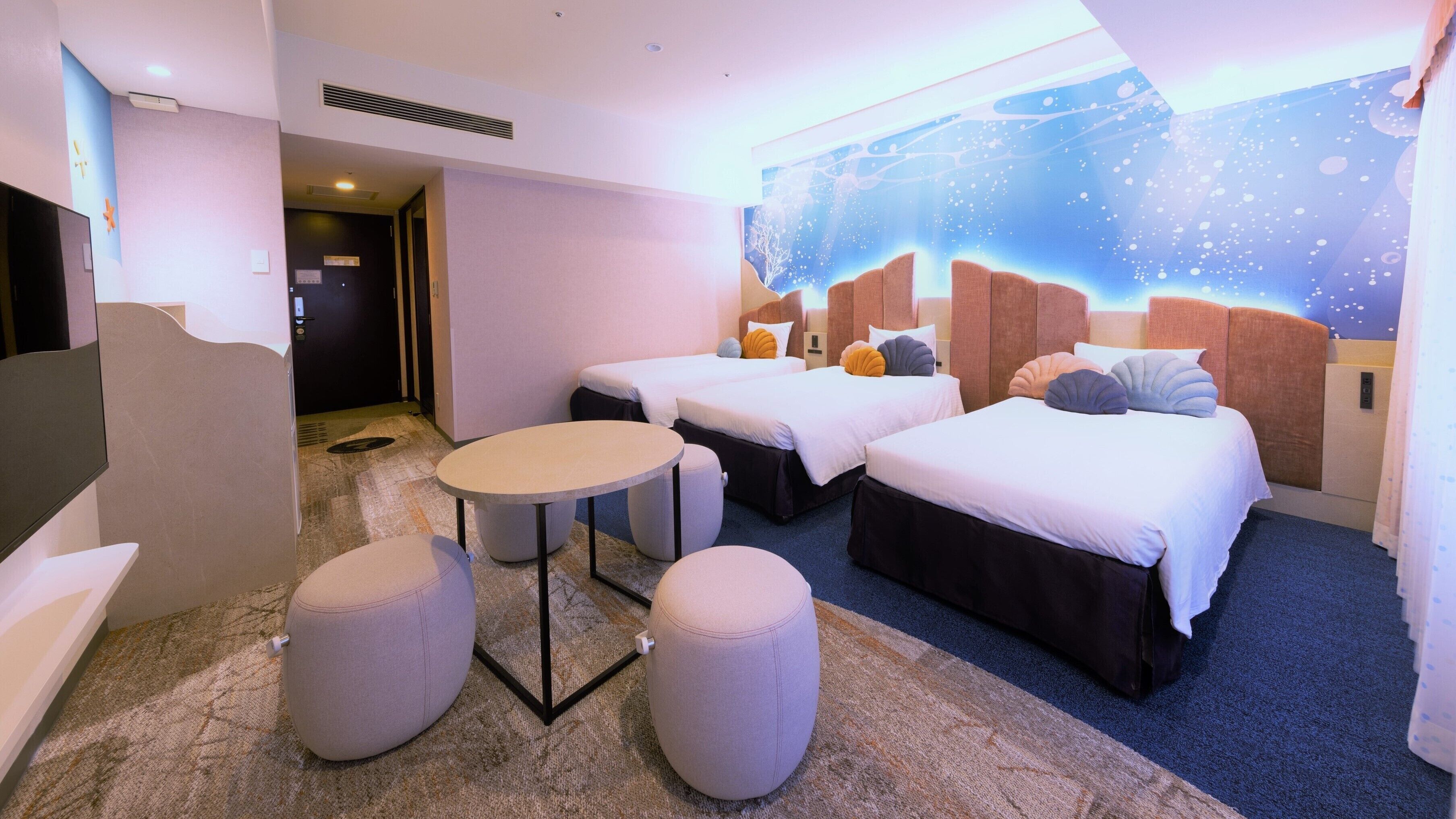 A hotel room with two beds, a small table, two ottomans, a TV, and a wall mural.