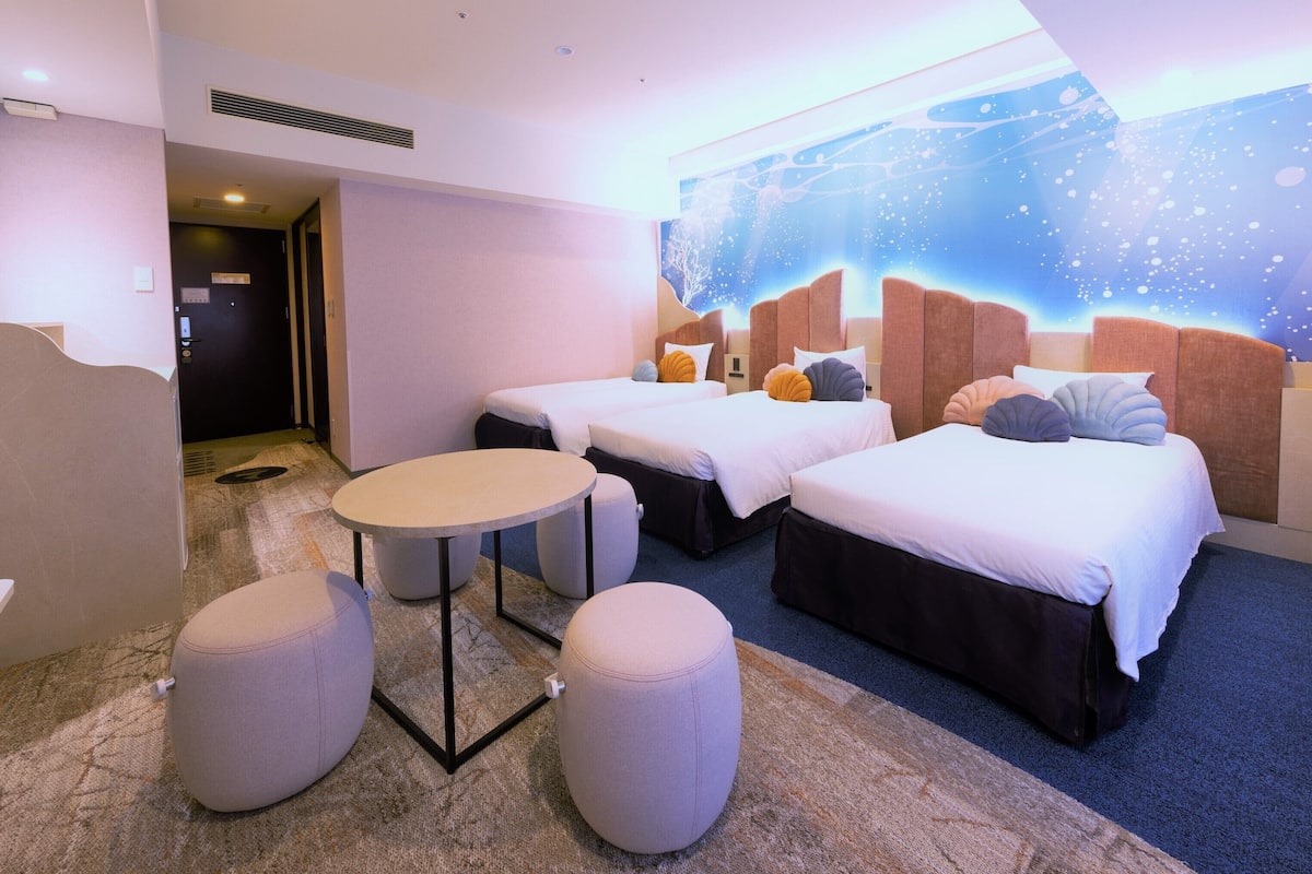 A hotel room with two beds, a small table, two ottomans, a TV, and a wall mural.