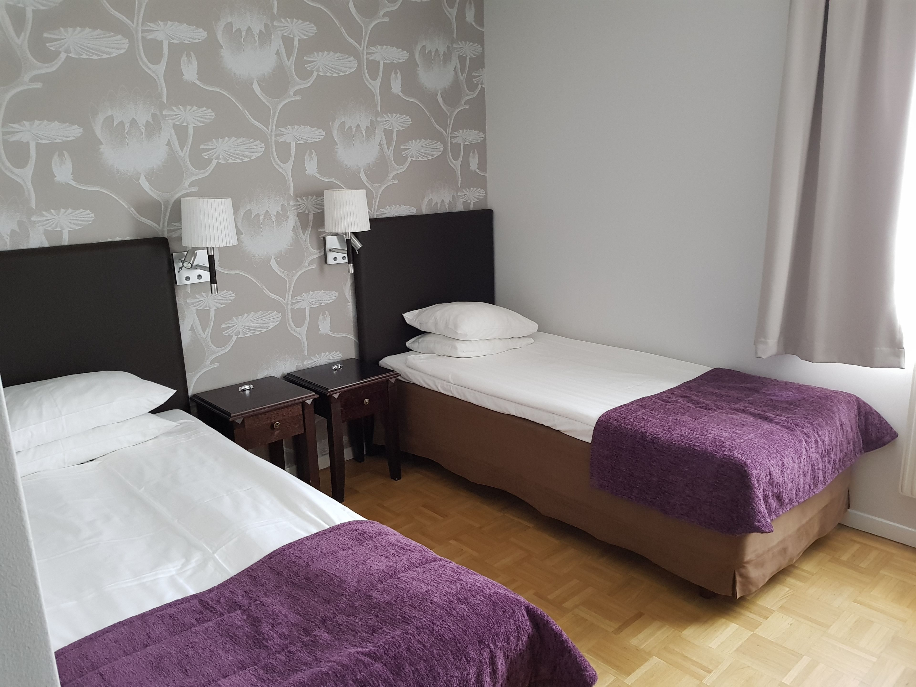 A hotel room with two single beds, a wooden floor, a floral wallpaper, and a window with curtains.