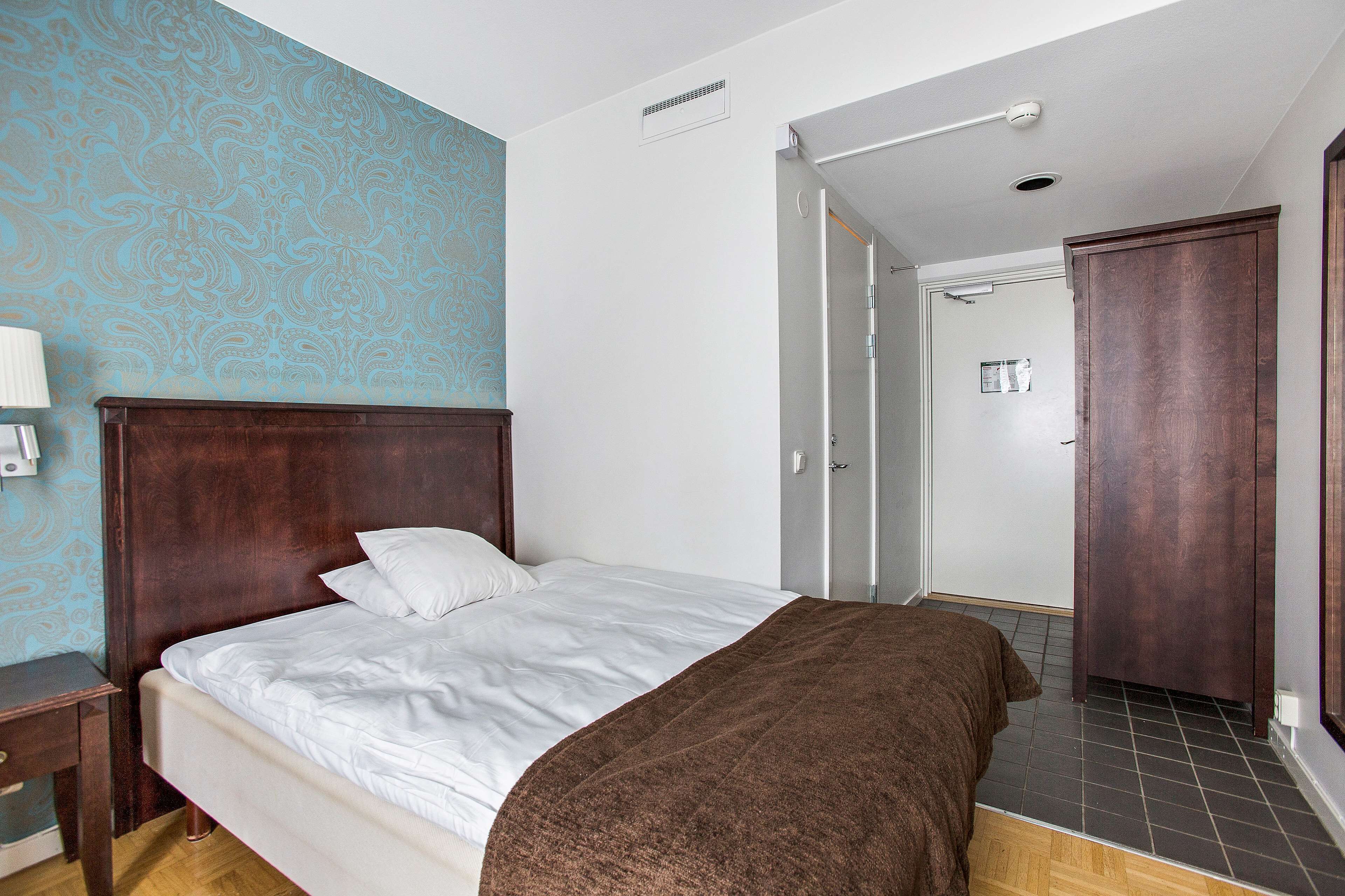 A hotel room with a wooden bed, white linens, a brown throw blanket, a nightstand with a lamp, and a patterned wall.