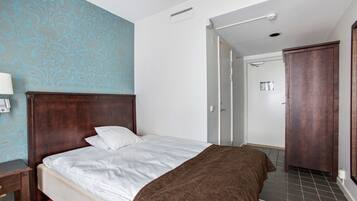 A hotel room with a wooden bed, white linens, a brown throw blanket, a nightstand with a lamp, and a patterned wall.