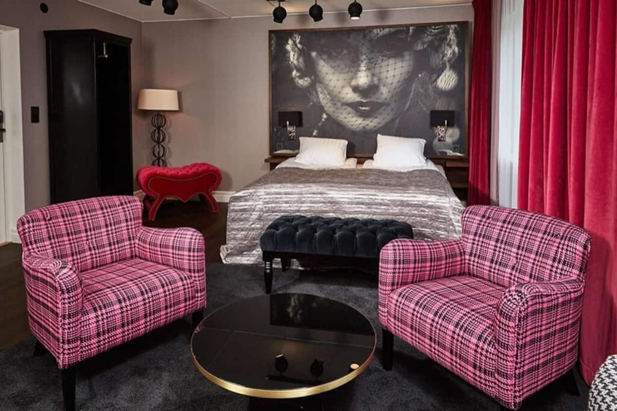 A bedroom with a bed, two armchairs, a round coffee table, a red heart-shaped ottoman, a black wardrobe, and a large wall art piece.
