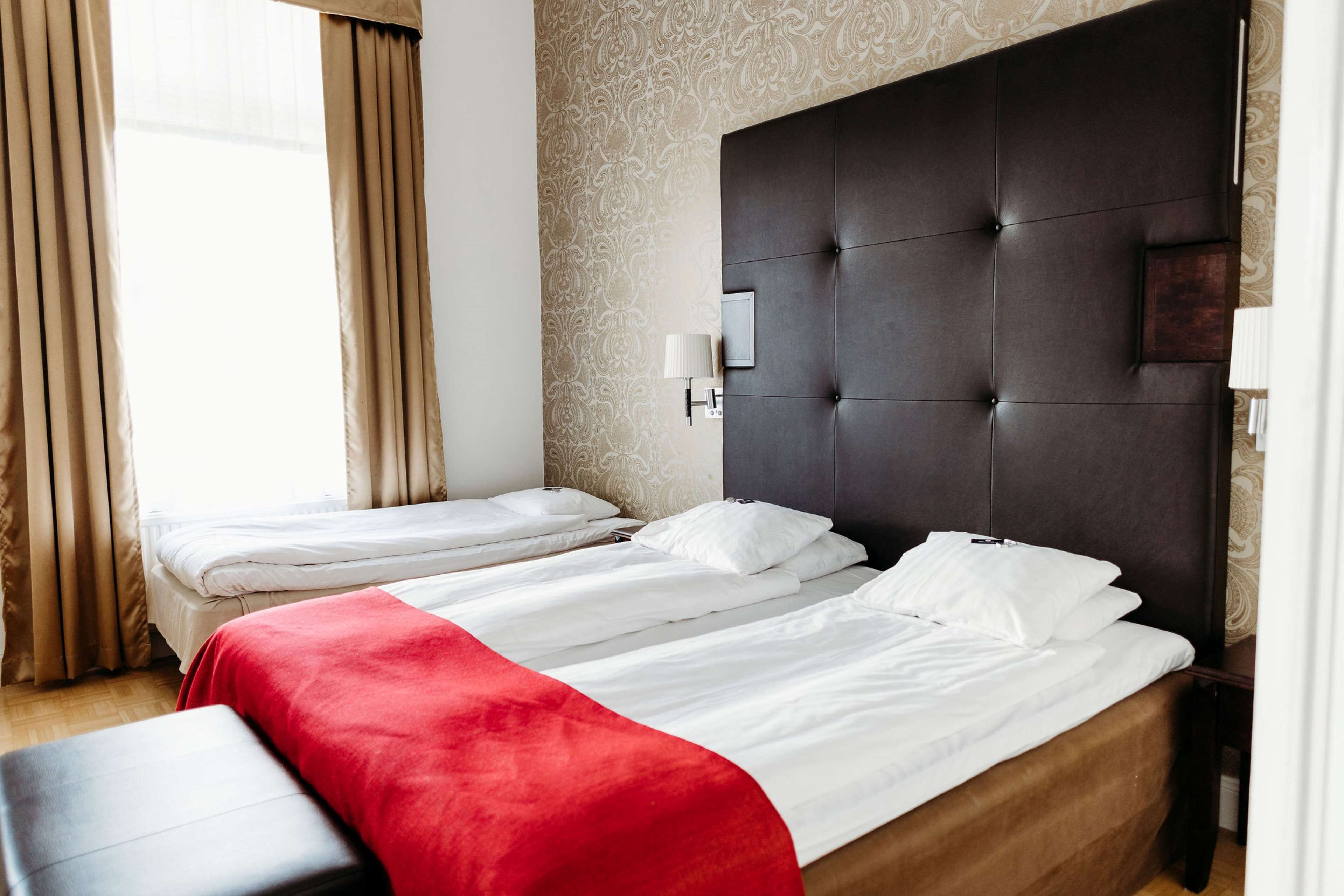 A hotel room with two single beds, a red throw blanket, a dark headboard, and a patterned wallpaper.