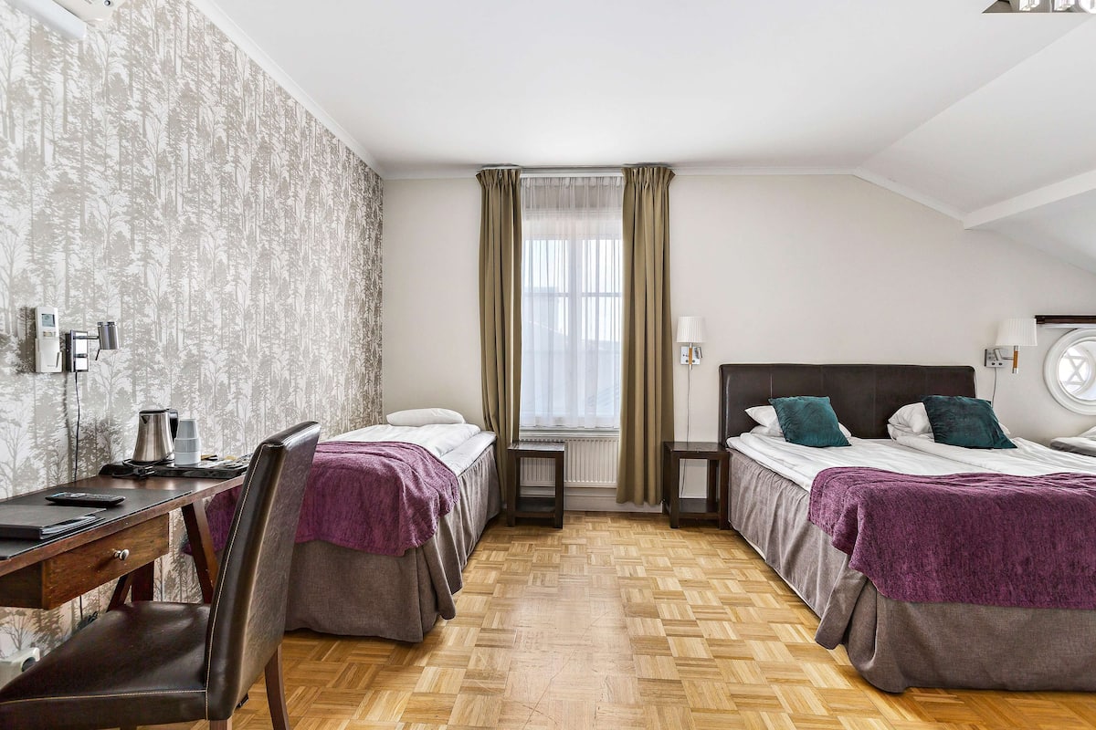 A hotel room with two beds, a desk, a chair, a radiator, and a window with curtains.