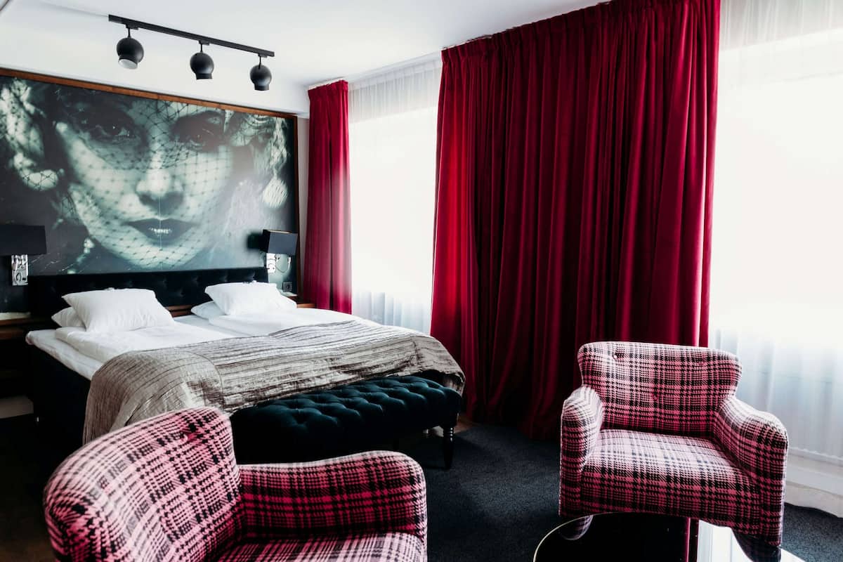 A hotel room with a large bed, two armchairs, a red curtain, and a wall-mounted TV.