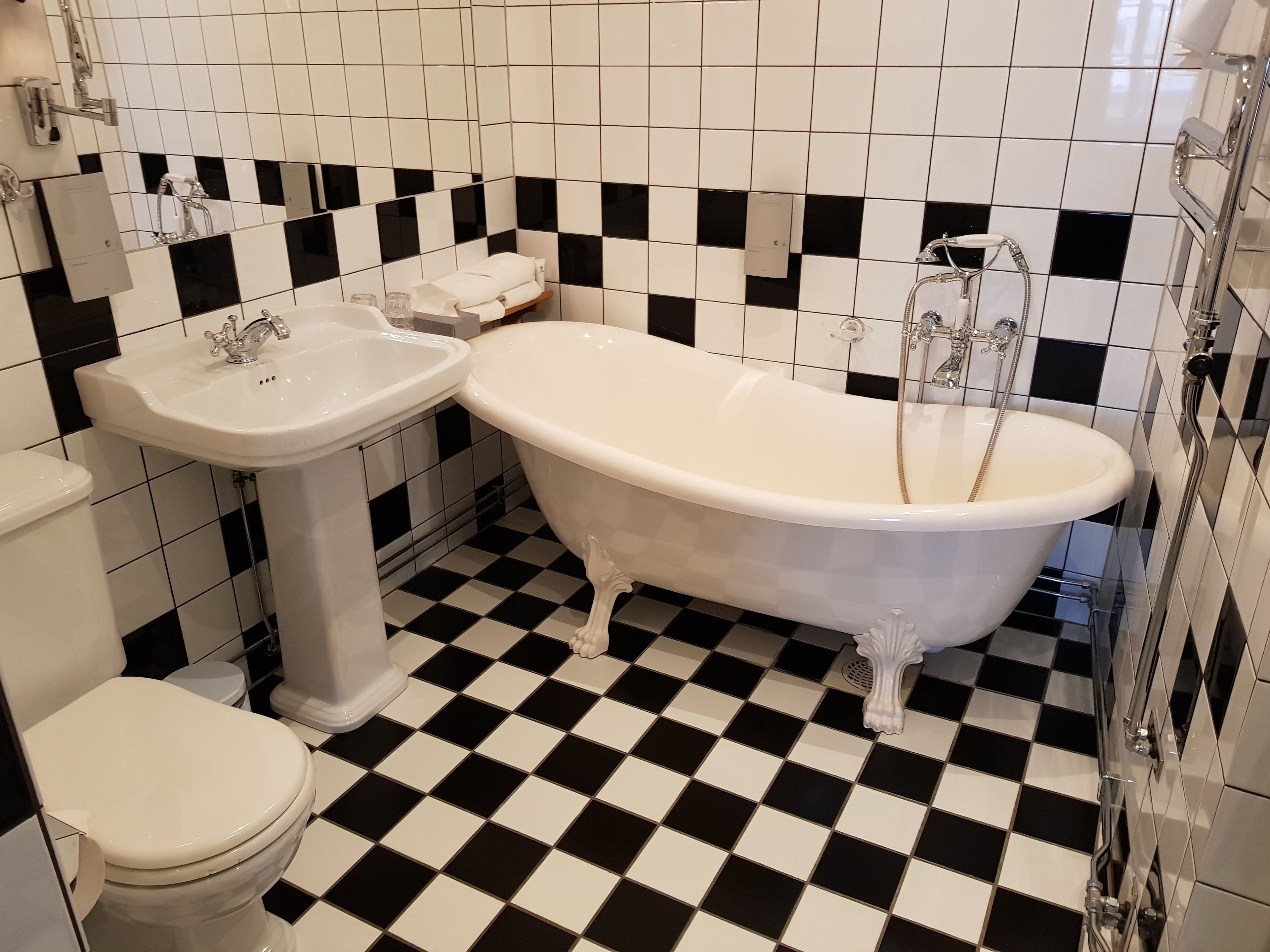 Ett badrum med ett svart- och vitrutigt golv, ett vitt handfat på en piedestal, ett badkar med tassar och en toalett.