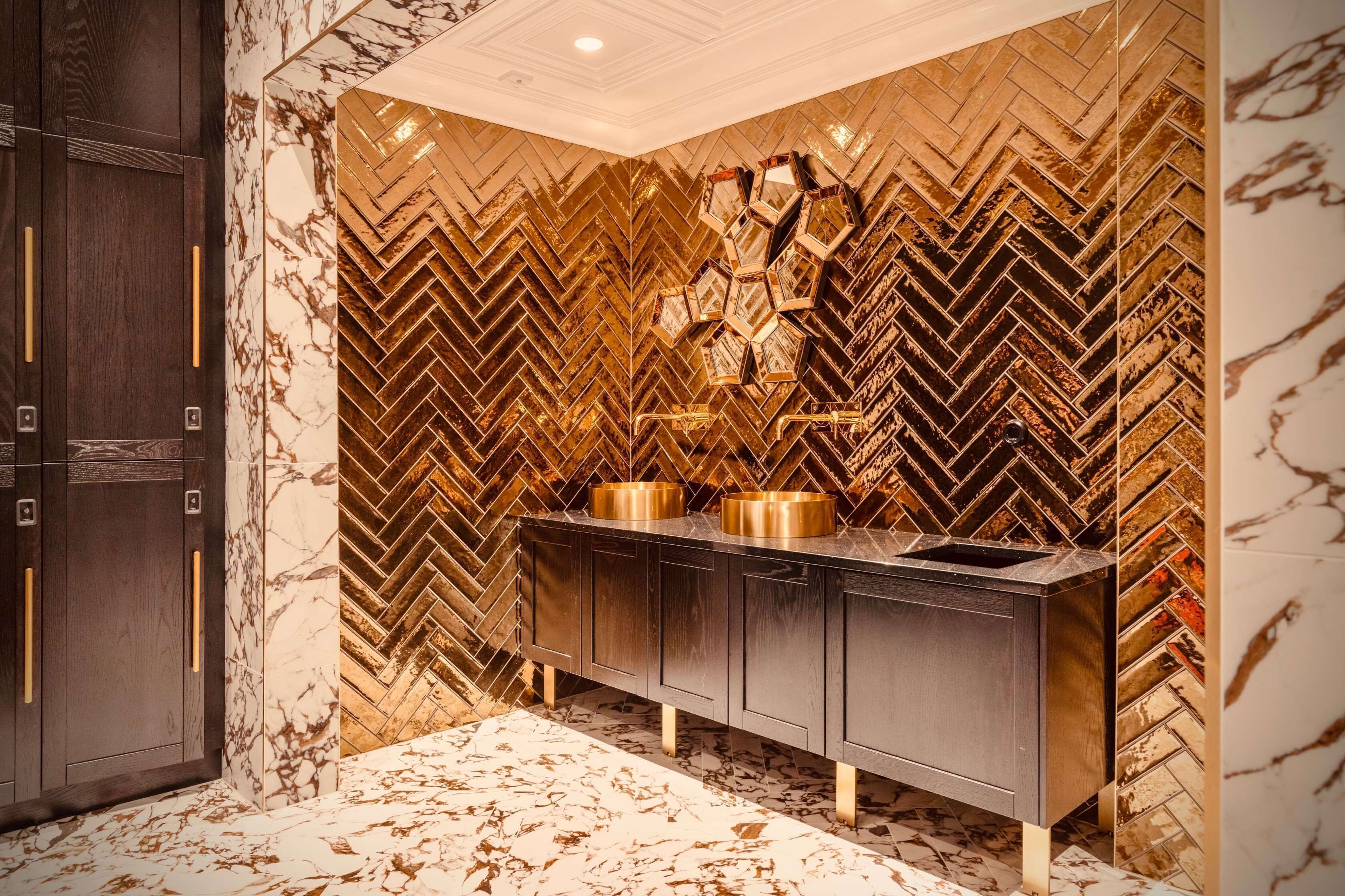 Un baño moderno con una pared de diseño geométrico tipo espiga, suelo de mármol y tocador de madera oscura con detalles dorados.