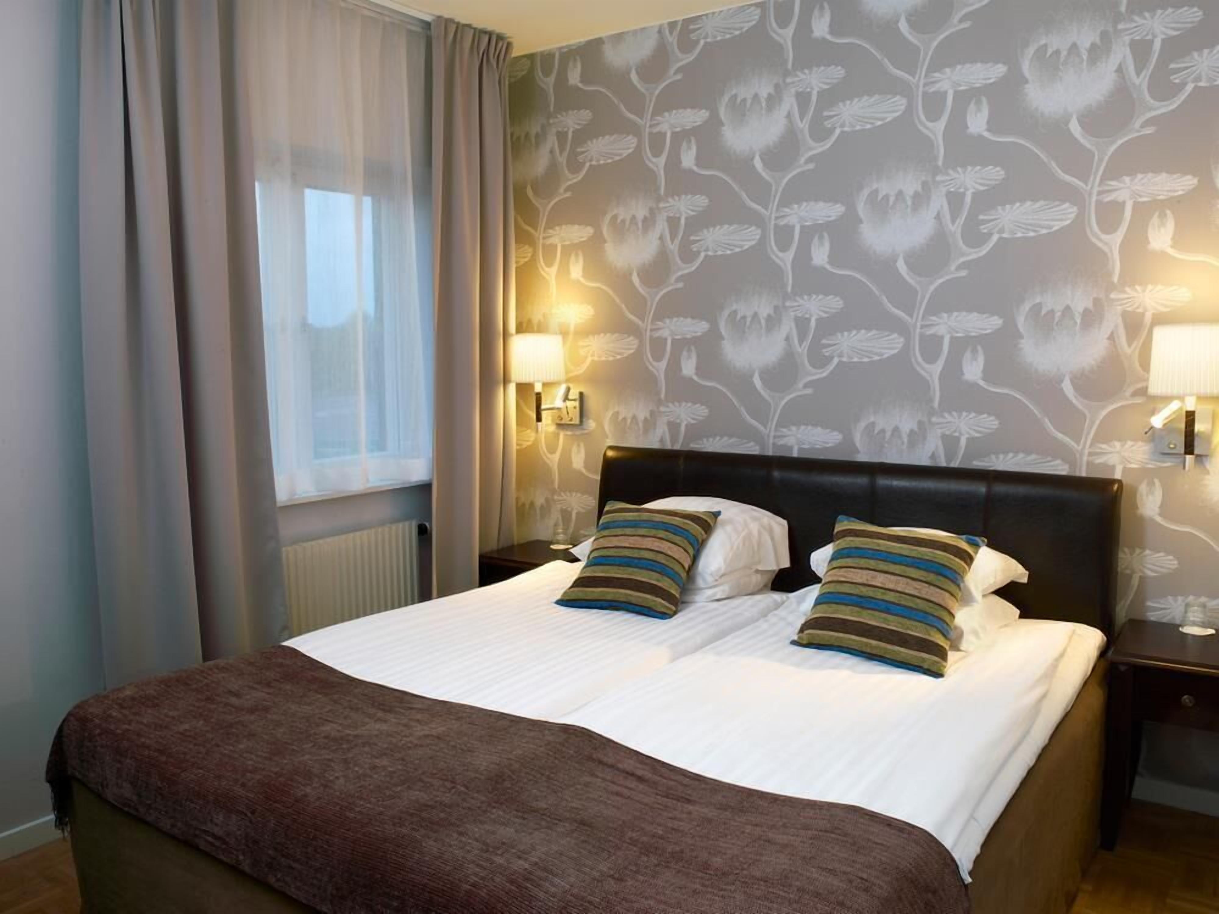 A neatly made bed with a brown blanket, two pillows, and a headboard. There are two bedside lamps and a wall with a floral pattern.