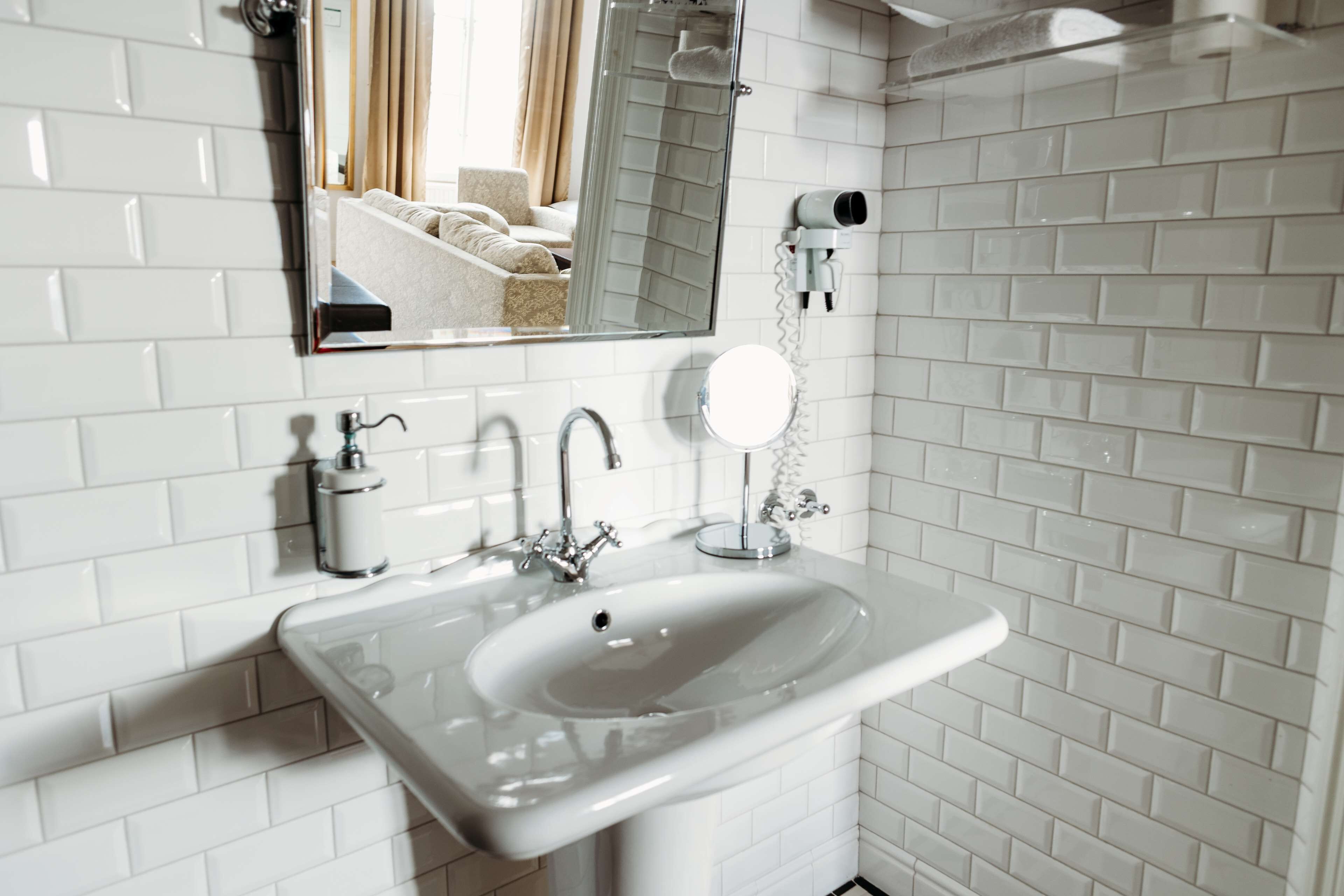 Un baño con paredes de azulejos blancos, un lavamanos blanco con un espejo encima y un secador de pelo en la pared, además de un espejo redondo pequeño sobre el lavamanos.