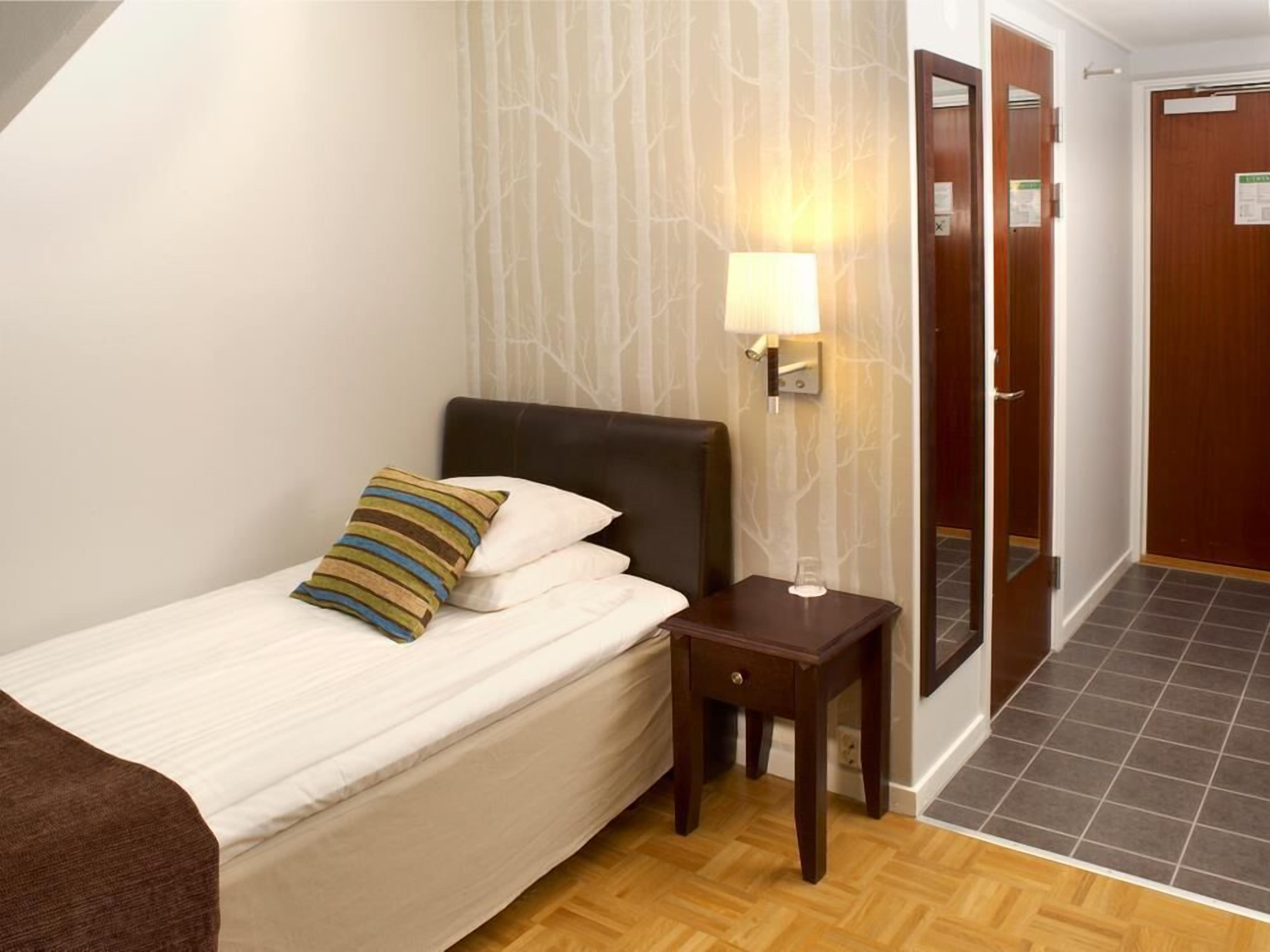 Una habitación de hotel con una cama, una mesita de noche, una lámpara y una puerta.