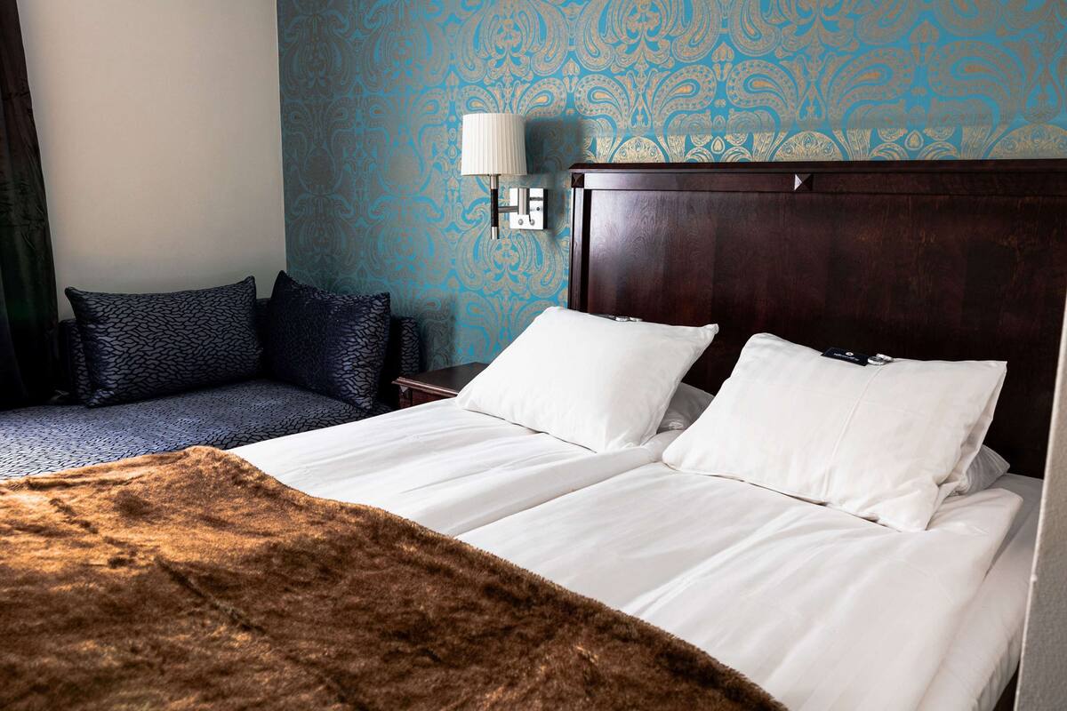 A hotel room with a large bed, a smaller sofa, a lamp, and a patterned wall.