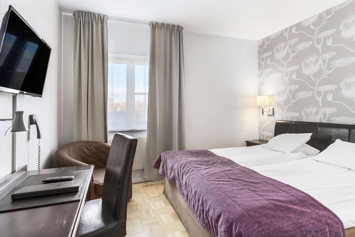 A hotel room with a bed, a desk with a computer, a chair, a TV, and a window with curtains.