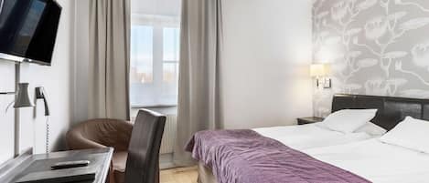 A hotel room with a bed, a desk with a computer, a chair, a TV, and a window with curtains.