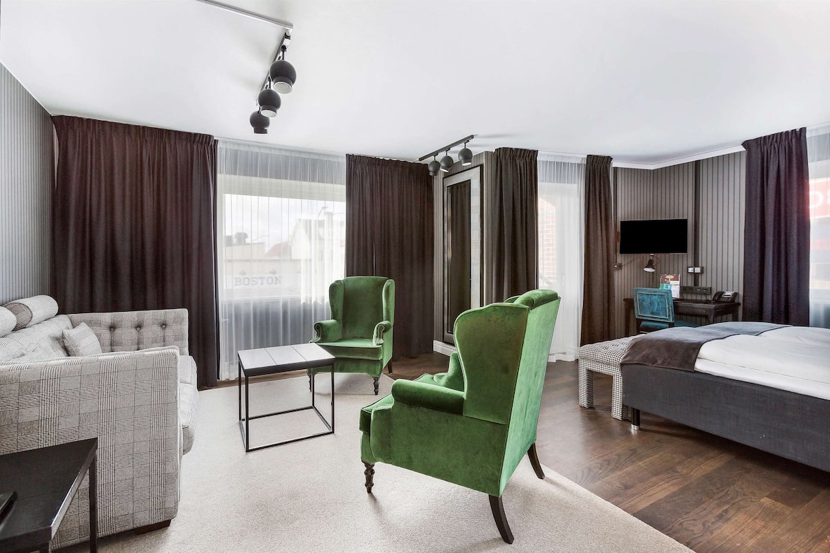 A hotel room with a sofa, a green armchair, a bed, a desk with a chair, a TV, and a window with curtains.