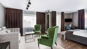 Ein Hotelzimmer mit einer Couch, einem grünen Sessel, einem Bett, einem Schreibtisch mit Stuhl, einem Fernseher und einem Fenster mit Vorhängen.