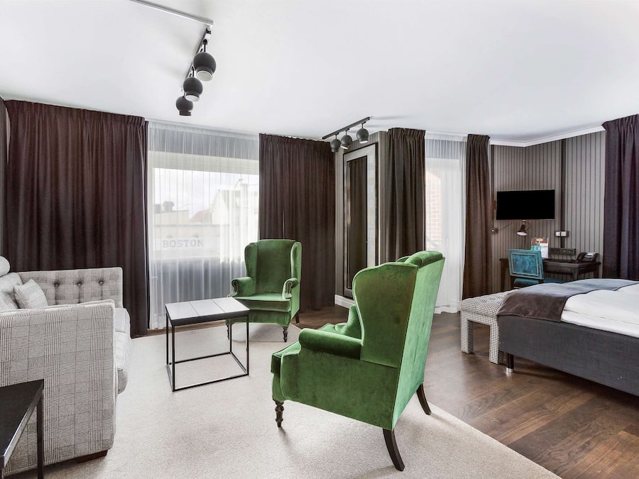 Ein Hotelzimmer mit einer Couch, einem grünen Sessel, einem Bett, einem Schreibtisch mit Stuhl, einem Fernseher und einem Fenster mit Vorhängen.