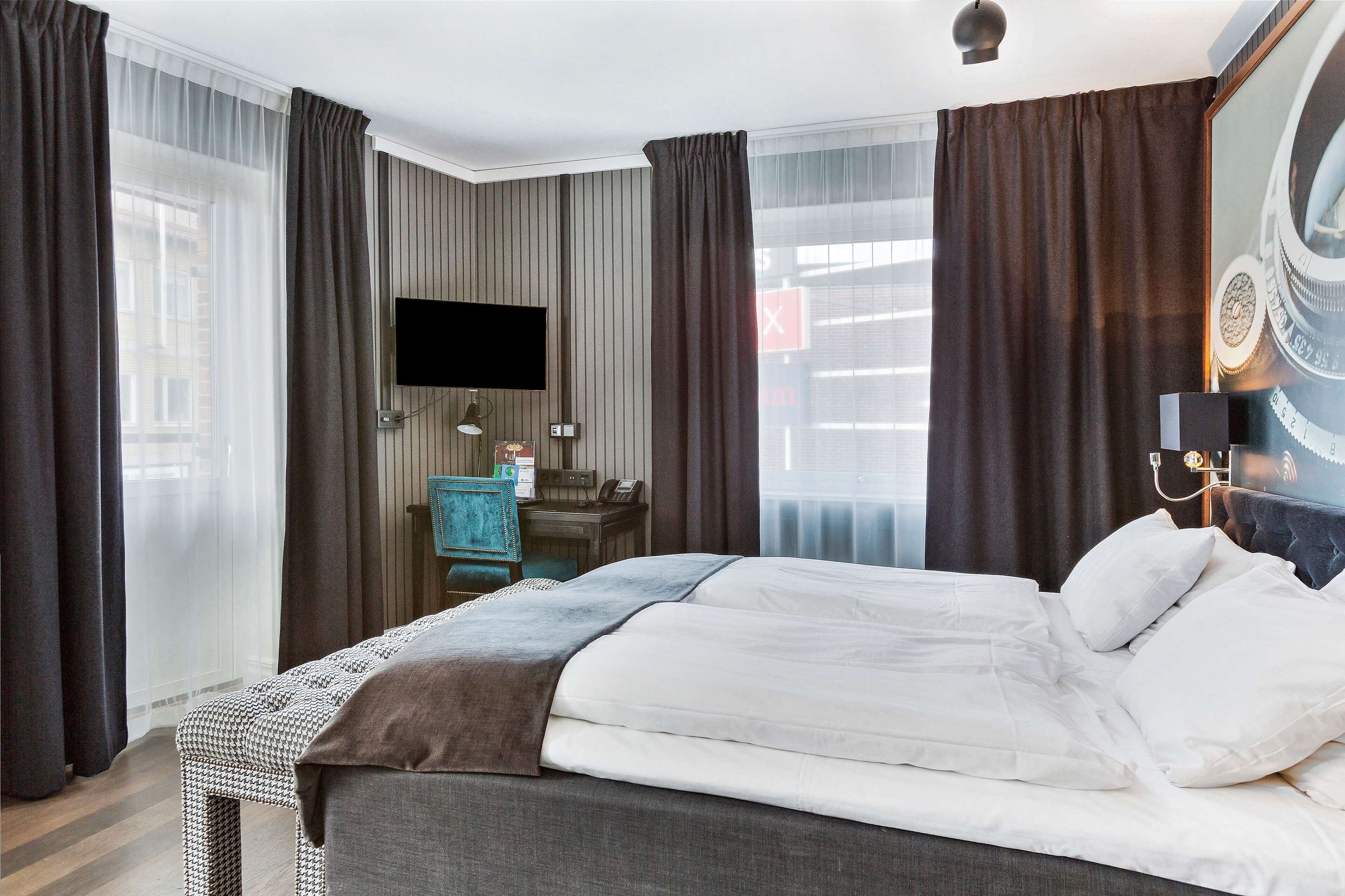 A hotel room with a large bed, a desk with a lamp, a TV, and a view of the city through the window.