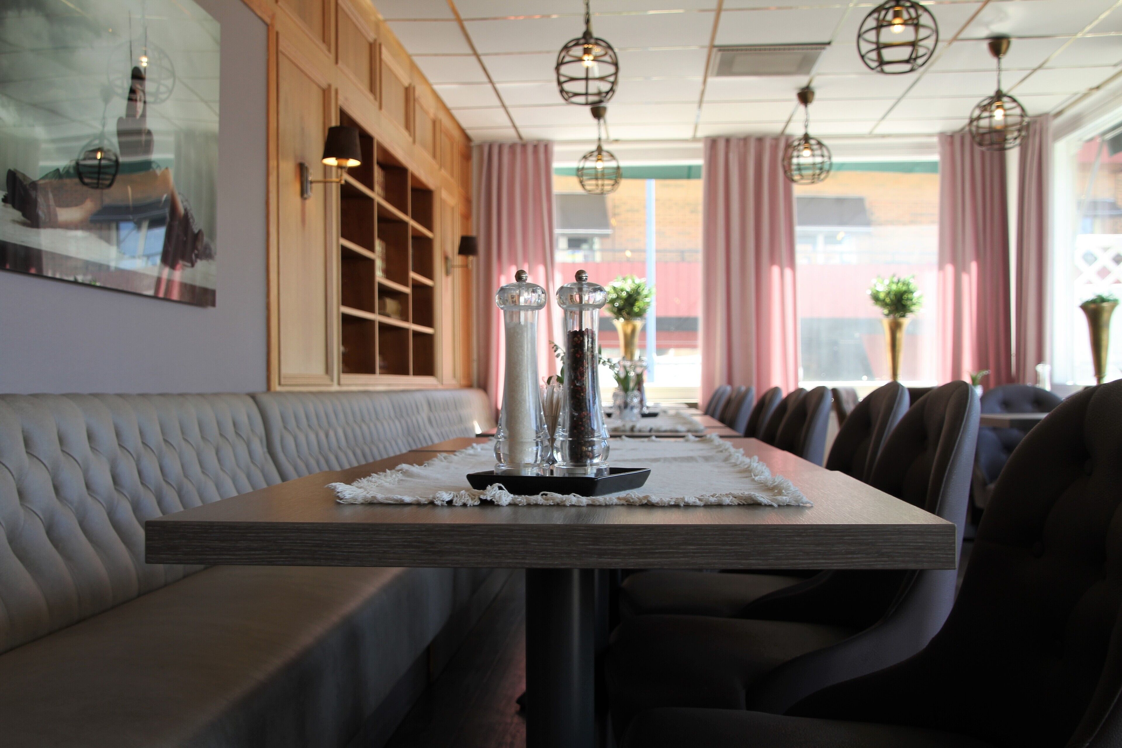Un restaurante con una larga mesa de madera, asientos acolchados, lámparas colgantes y vistas amplias a través de grandes ventanales.