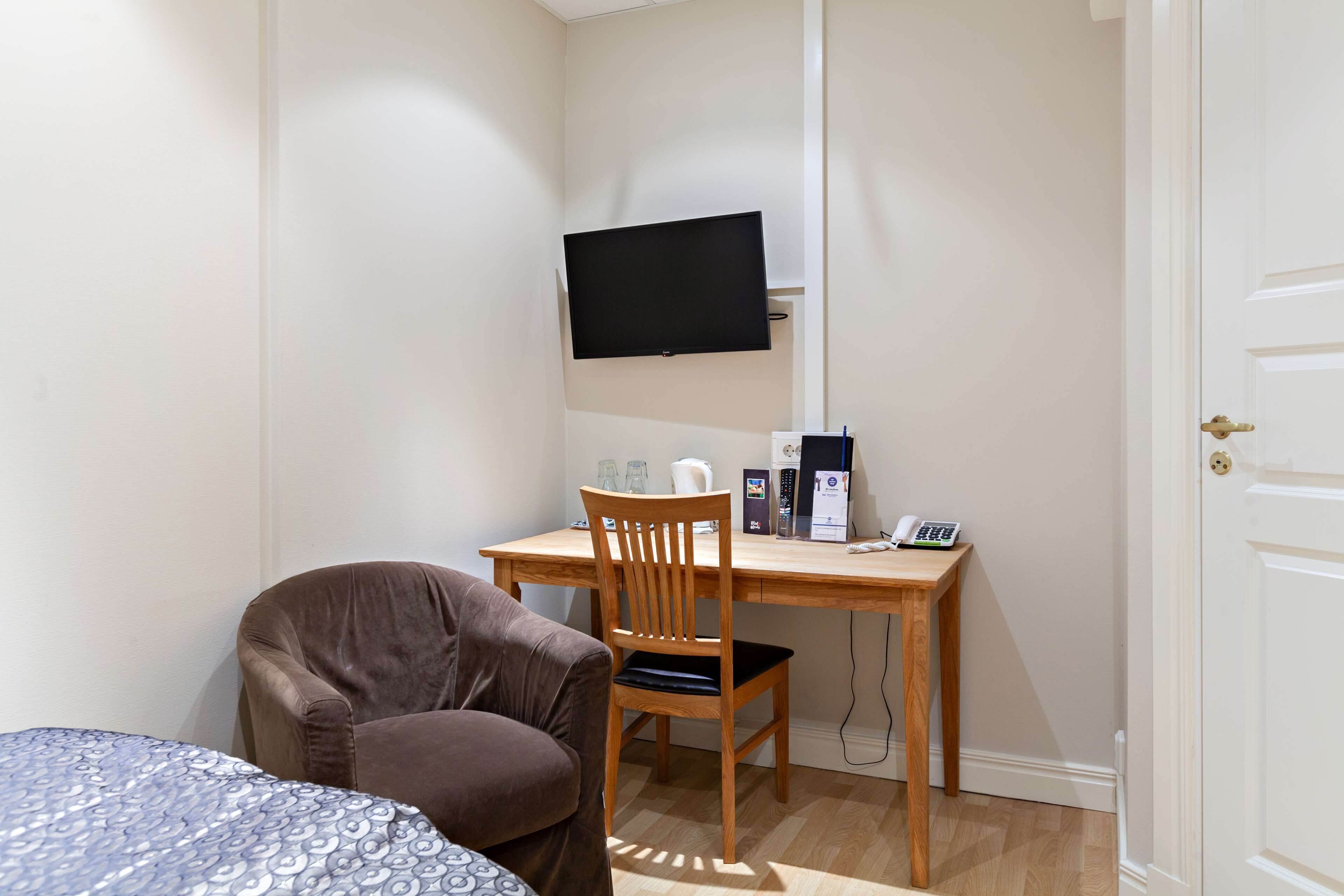 Een kleine kamer met een bed, een bureau met stoel, een aan de muur bevestigde televisie en een deur.