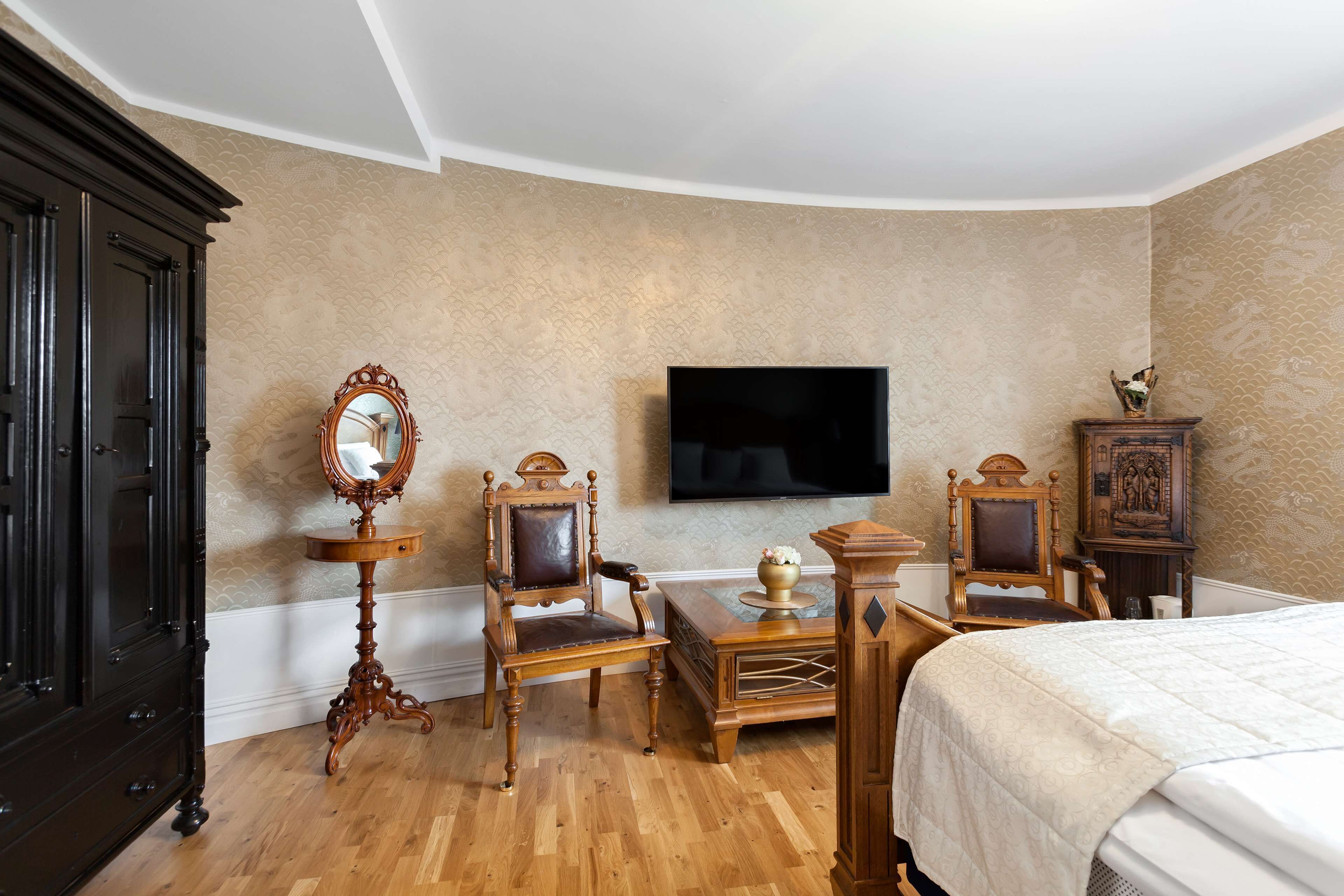 A room with a bed, a television mounted on the wall, a wooden cabinet, and a chair with a mirror.