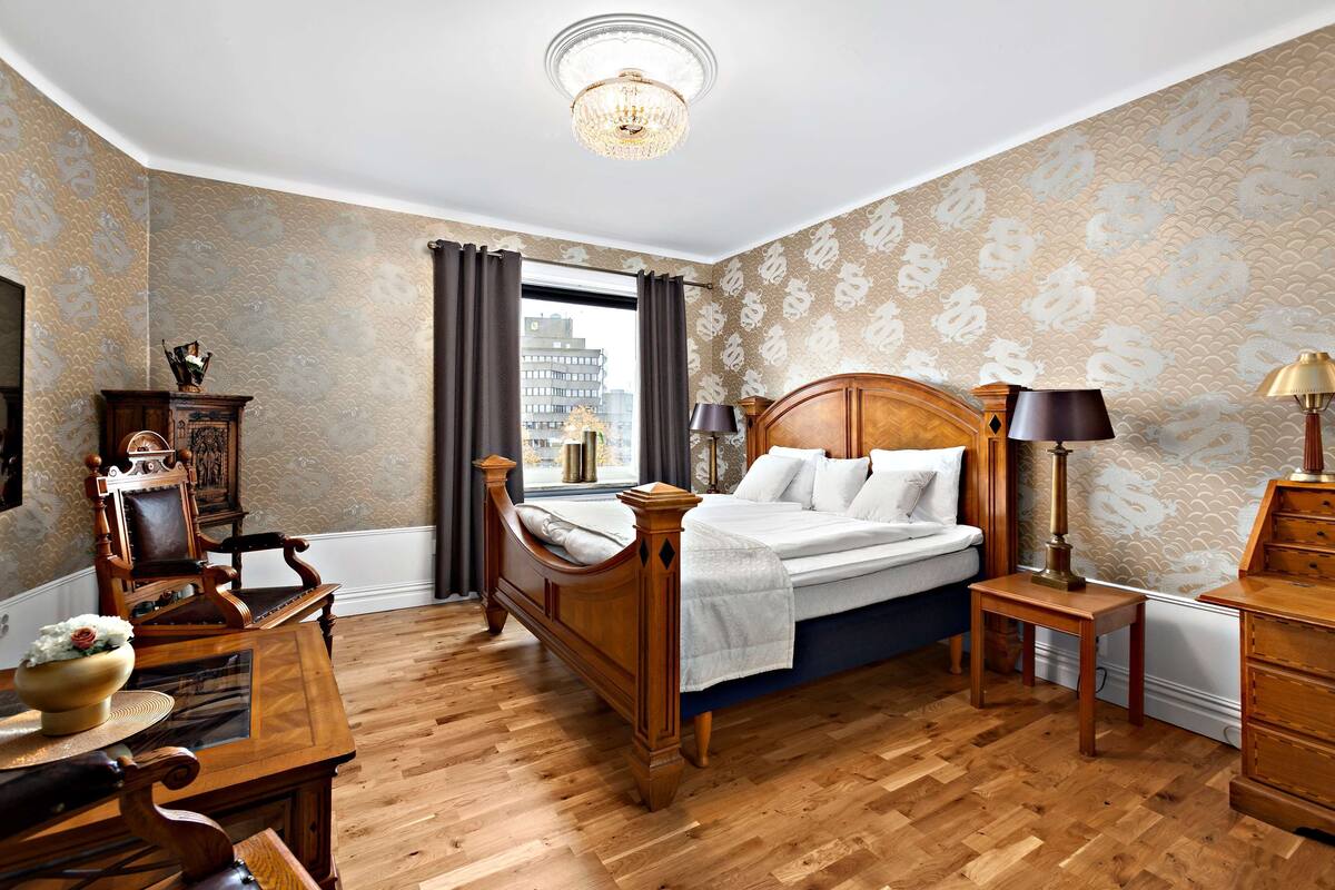A hotel room with a wooden bed, a desk, a chair, a lamp, and a view of the city through the window.