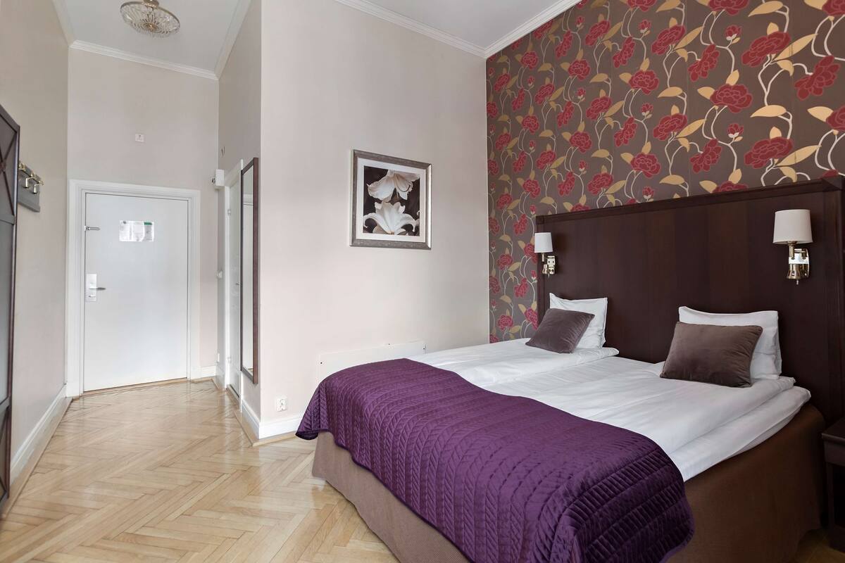 A hotel room with a bed, a purple blanket, a floral wallpaper, a framed picture on the wall, and a wooden floor.