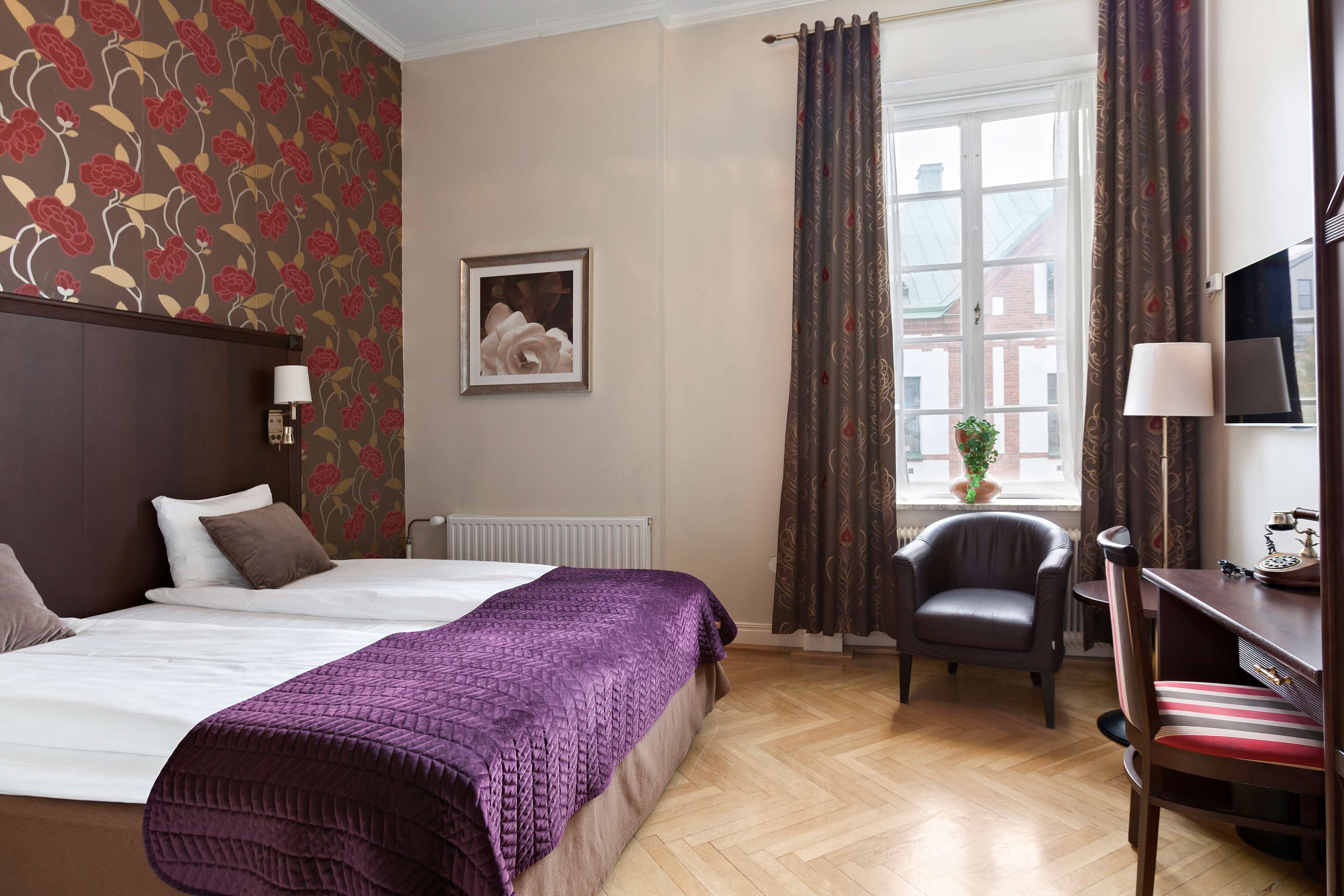 Hotelkamer met een bed, bureau, stoel, raam met gordijnen, radiator en een ingelijst schilderij aan de muur.