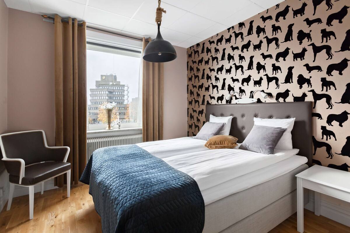 A hotel room with a bed, a chair, a nightstand, and a window with curtains.