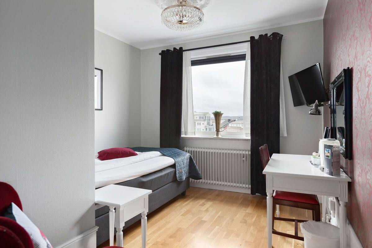 A small, well-lit room with a bed, a desk, and a window with curtains.