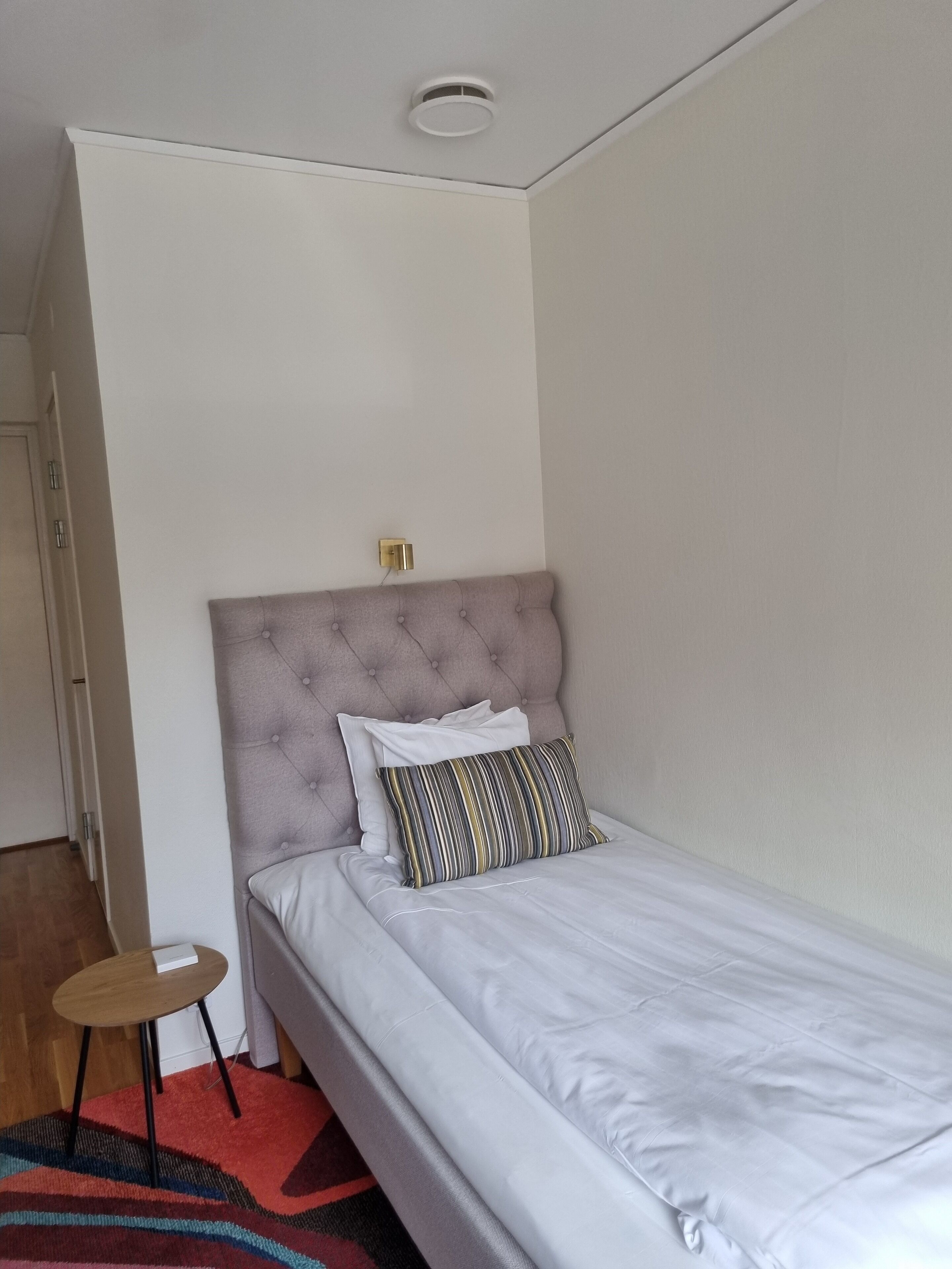 A single bed with a tufted headboard, a bedside table, and a power outlet on the wall.