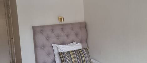 A single bed with a tufted headboard, a bedside table, and a power outlet on the wall.