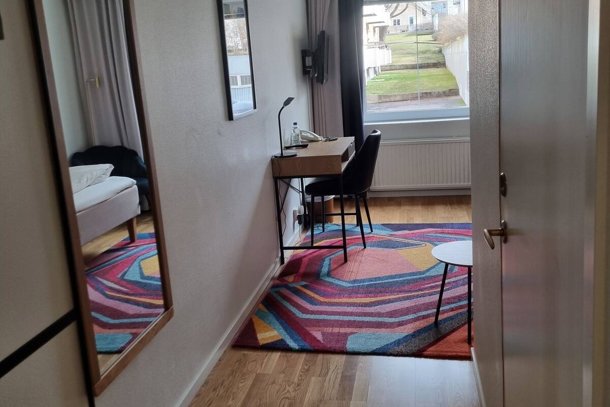 A hotel room with a wooden floor, a desk with a chair, a mirror, a bed, and a window with curtains.