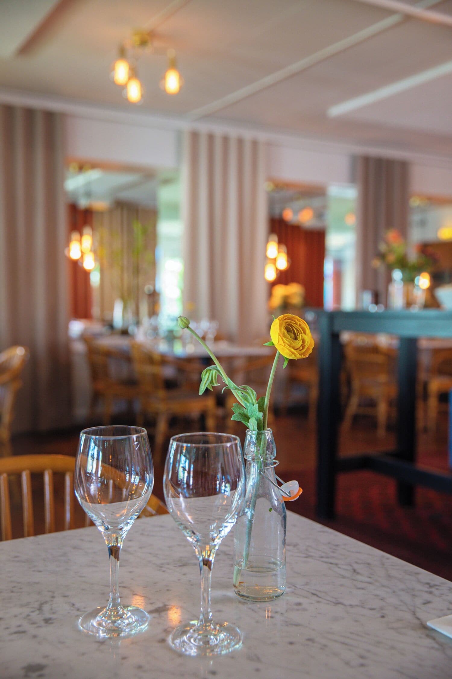 A restaurant setting with a marble table, two empty wine glasses, and a vase with a single yellow flower.